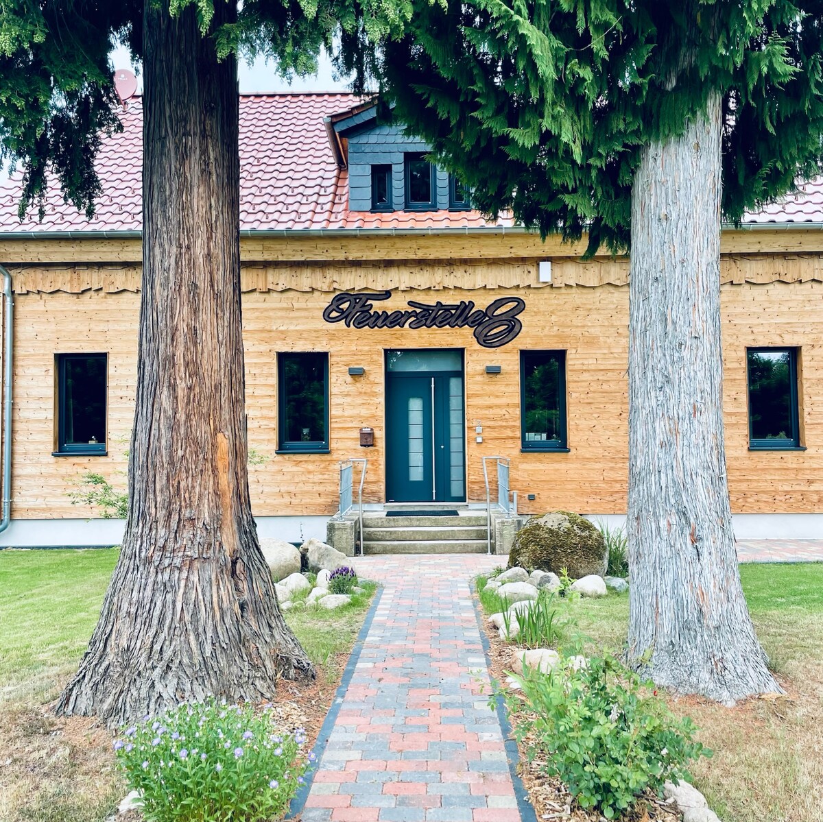 An inviting entrance to Fuerstelle8 is framed by tall trees. The building features natural wood siding and a sleek door outlined by decorative lighting. A stone pathway leads to the entrance, surrounded by manicured grass and a few decorative stones.