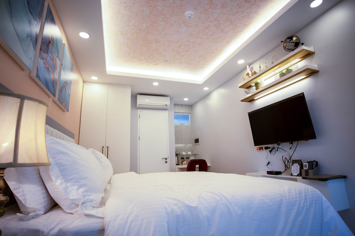 A well-lit room features a spacious bed with neatly arranged white linens. A flat-screen TV is mounted on a wall over a console. In the background, a small bathroom area is visible, complemented by modern cabinetry and decorative accents.