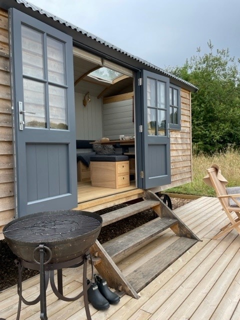 The entrance of a charming shepherd's hut is framed by double doors, revealing a cozy interior with seating and wooden accents. Steps lead down to a deck, where a circular fire pit and a pair of welly boots are positioned. Surrounding greenery enhances the rustic setting.