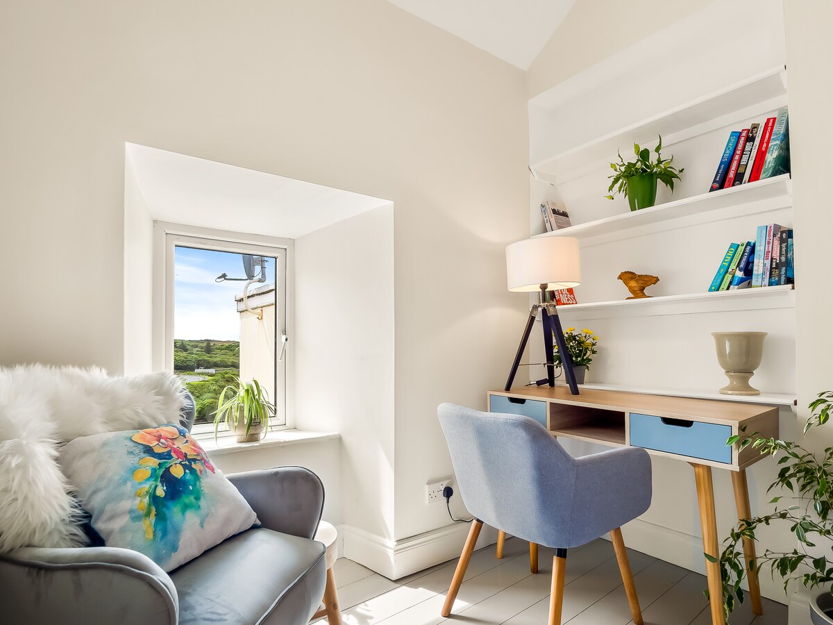 A bright corner workspace features a small desk with a light blue front, accompanied by a chair and a lamp. A comfortable armchair with decorative cushion is positioned nearby, and a window offers a view of greenery outside.