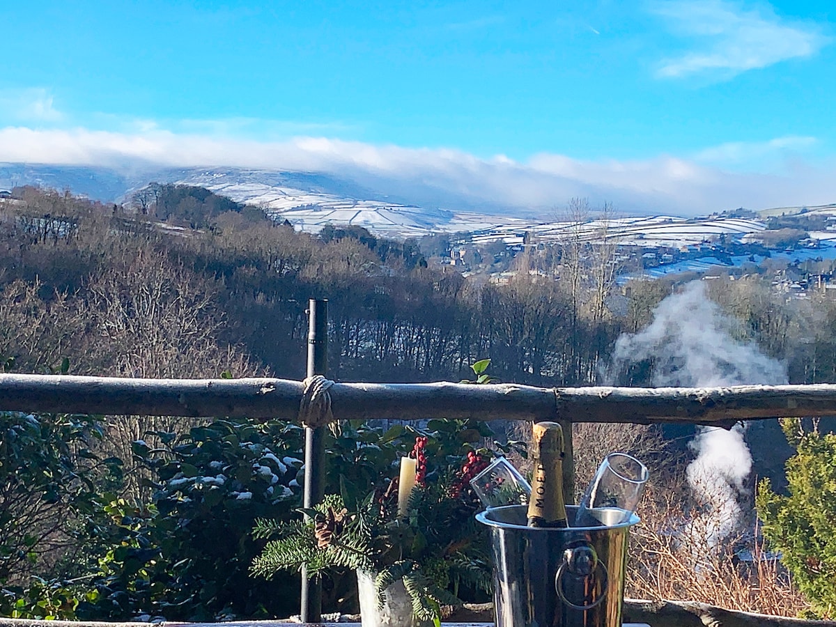 A view of the snow-covered hills is framed by a rustic wooden railing. A bucket of champagne sits in the foreground alongside two fluted glasses, with wisps of steam visible in the distance, hinting at the crisp winter air.