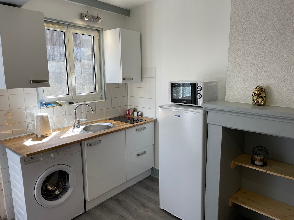 A well-equipped kitchen is showcased, featuring modern white cabinetry and a wooden countertop. A washing machine and refrigerator are visible, along with a microwave. Natural light enters through a window, illuminating the space. Open shelving is positioned beside the kitchen area.