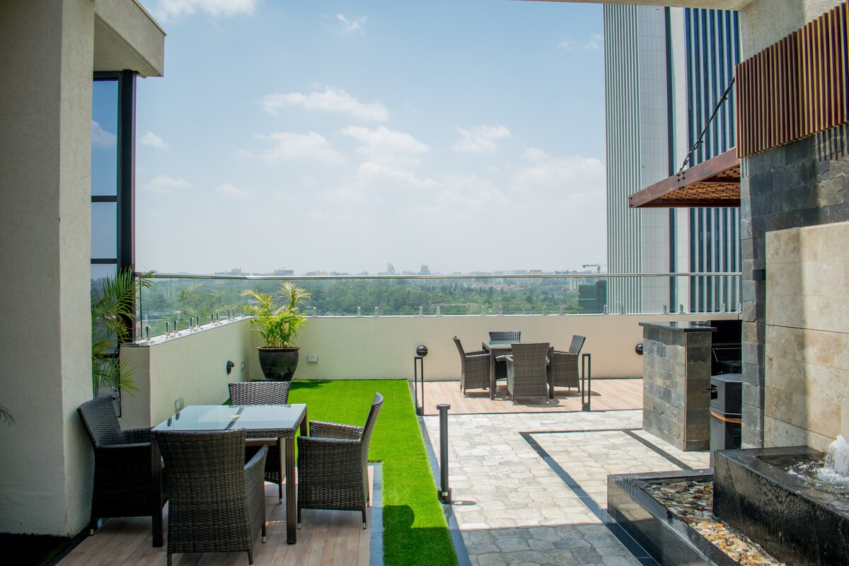 A spacious rooftop terrace features comfortable seating arrangements with wicker chairs and tables. Lush green grass accents the area, which is complemented by stone flooring. A barbecue grill is visible, offering inviting views of the surrounding landscape.