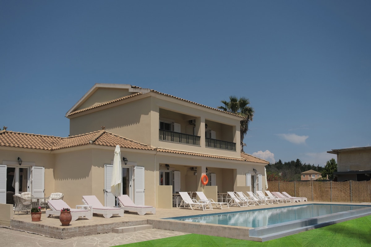 A spacious, two-story building with a traditional design is shown beside a newly constructed pool. Sun loungers and umbrellas are arranged around the pool area, and palm trees create a relaxed ambiance. The building features multiple balconies with white shutters, reflecting a bright and inviting environment.