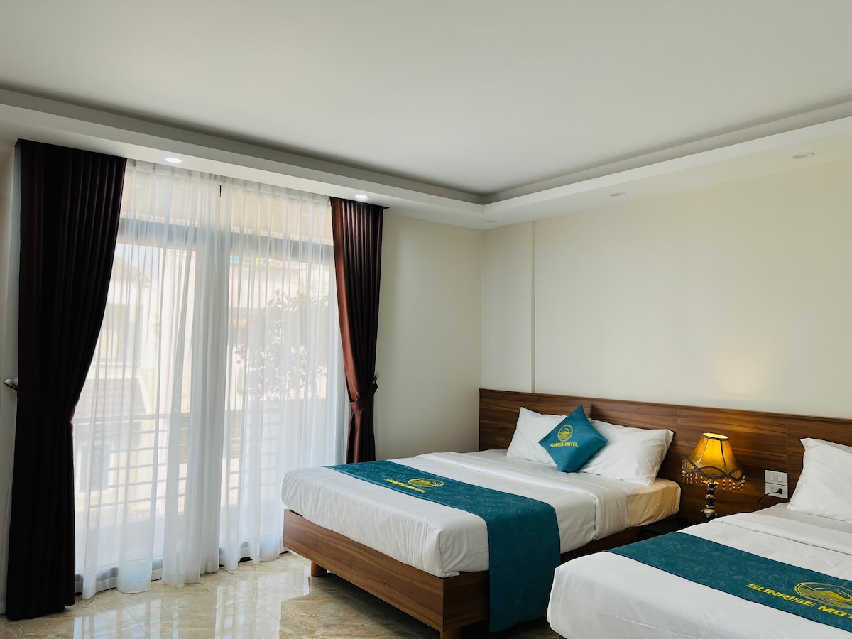 A bright sleeping area is highlighted by two spacious beds, each adorned with teal bed runners. Natural light filters through sheer curtains, illuminating the room's neutral tones and enhancing the inviting atmosphere. A bedside lamp adds a warm touch.