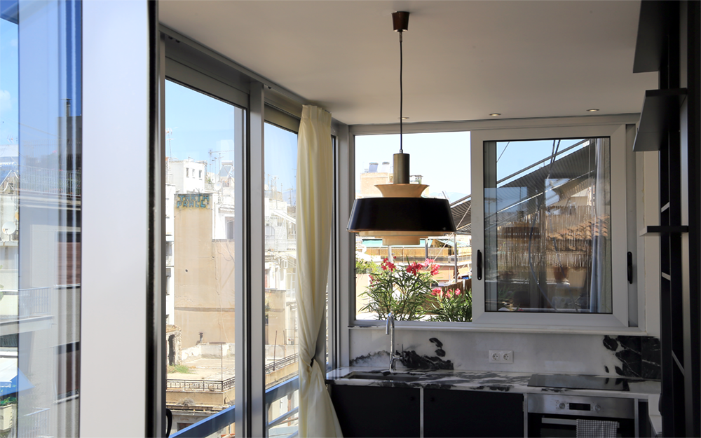 A well-lit kitchen area features large windows that offer views of the surrounding Athenian architecture. A modern pendant light hangs above the countertop, which reflects a stylish design with dark stone elements. A potted plant adds a touch of greenery to the space.