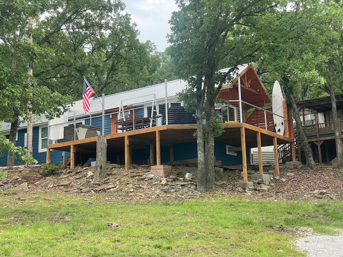 The exterior of the lake house features a wraparound deck elevated on sturdy posts, surrounded by greenery and natural stone. An American flag gently waves above the deck, which is adorned with seating for outdoor relaxation.