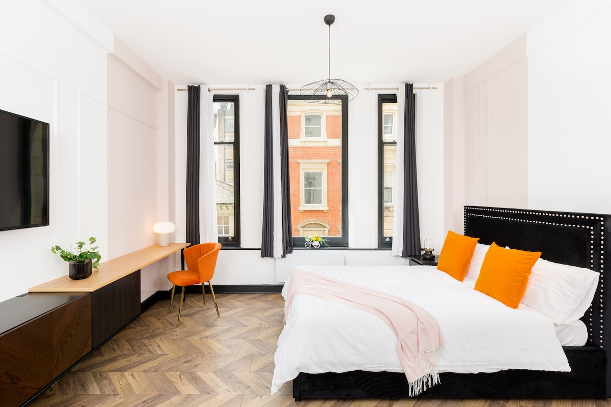 A stylish studio features a well-lit sleeping area with a spacious bed adorned with orange accents. A wooden desk and an orange chair provide a functional workspace. Tall windows allow natural light to fill the room, while a decorative light fixture adds a modern touch.
