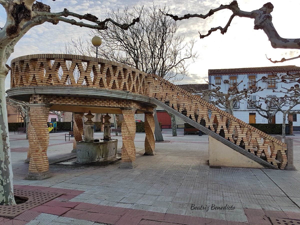 An artistic structure is displayed, featuring a sloped ramp and brickwork. A fountain is situated at the base, surrounded by flat paved areas. Leafless trees stand nearby, creating a contrast against the clear sky. Simple benches are arranged along the edges of the plaza.