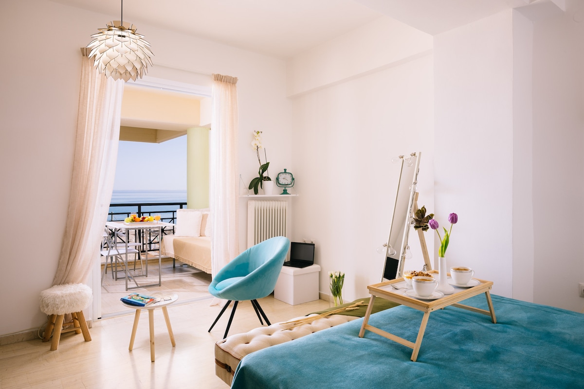 A bright and airy studio space features a double bed covered in a soft turquoise blanket. A small table with two cups and a vase of flowers is positioned near the bed. A seating area is visible through the open doors, overlooking the balcony with a view of the sea.