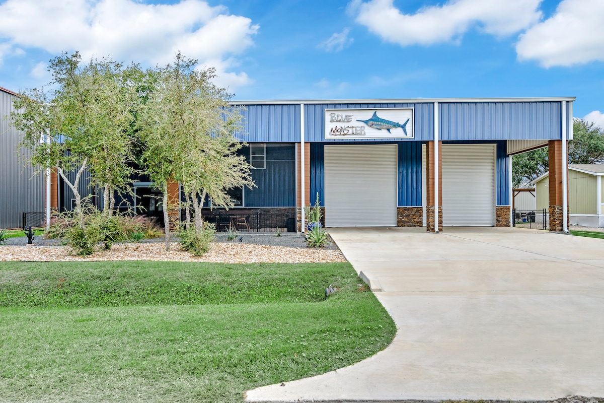 The exterior of the property features a large building with a metal roof and a welcoming sign reading 'Blue Monster.' A well-maintained lawn surrounds the structure, complemented by landscaped pebbles and a concrete driveway leading up to the entrance.