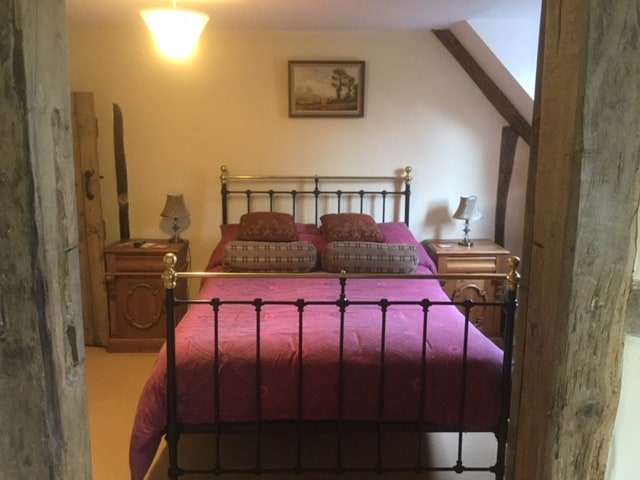 A spacious bedroom is presented, featuring a brass and iron bed adorned with decorative pillows. Nightstands are placed on either side, each with a lamp. Exposed wooden beams add character to the room, and a framed landscape painting is mounted above the bed.