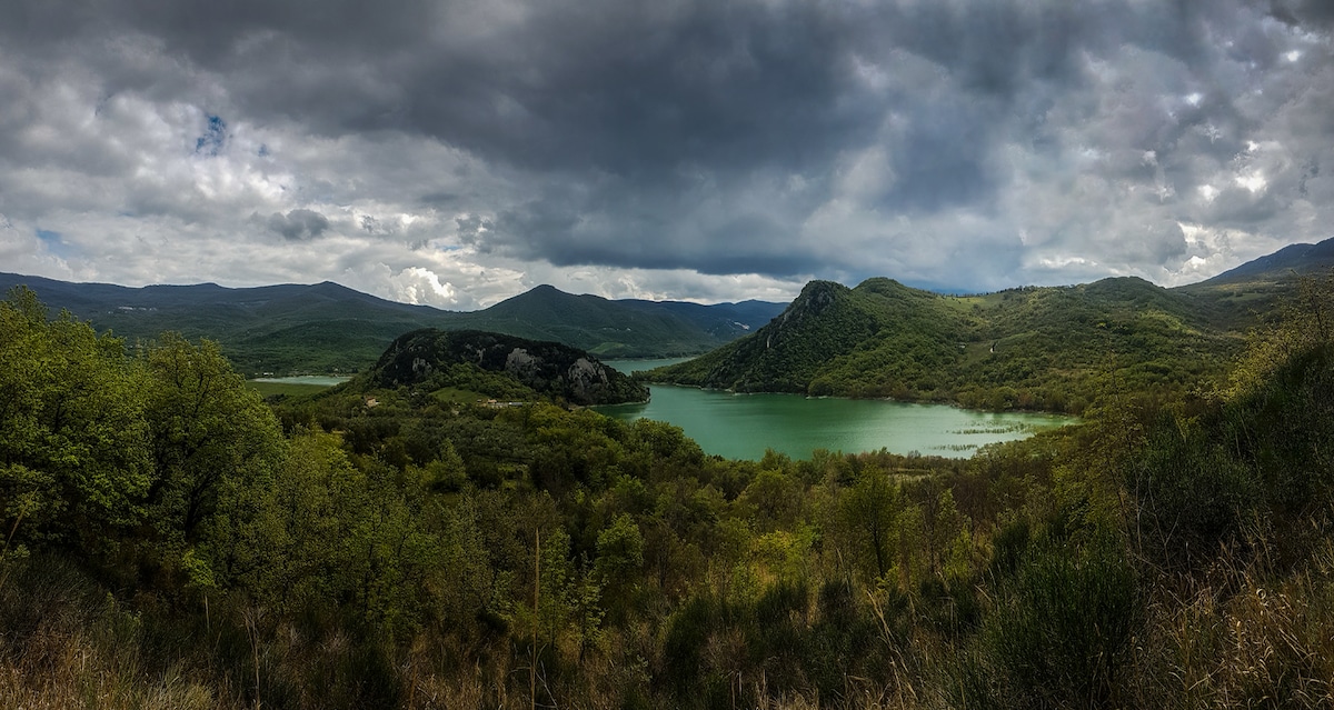A panoramic view of a serene lake surrounded by lush greenery and rolling hills. Dark clouds hover above the mountains in the distance, reflecting the tranquil yet dynamic landscape of Abruzzo. The rich colors of the water contrast with the vibrant foliage, creating an inviting natural setting.