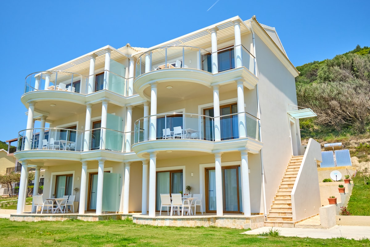 The three-story building features modern architectural design, with spacious balconies accessible from each suite. Sunlight reflects off the light-coloured exterior walls, and well-maintained grassy areas are visible in the foreground, providing a pleasant environment near the beach.
