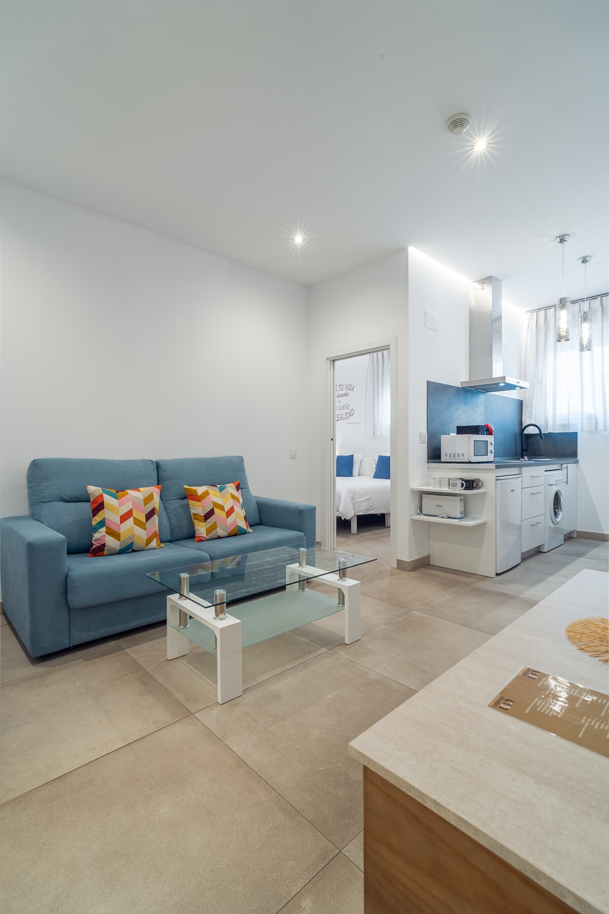 A living area features a light blue sofa adorned with patterned cushions, complemented by a glass coffee table. A well-equipped kitchenette is visible in the background, alongside a door leading to a bedroom. Soft lighting enhances the neutral tones of the tiled flooring.