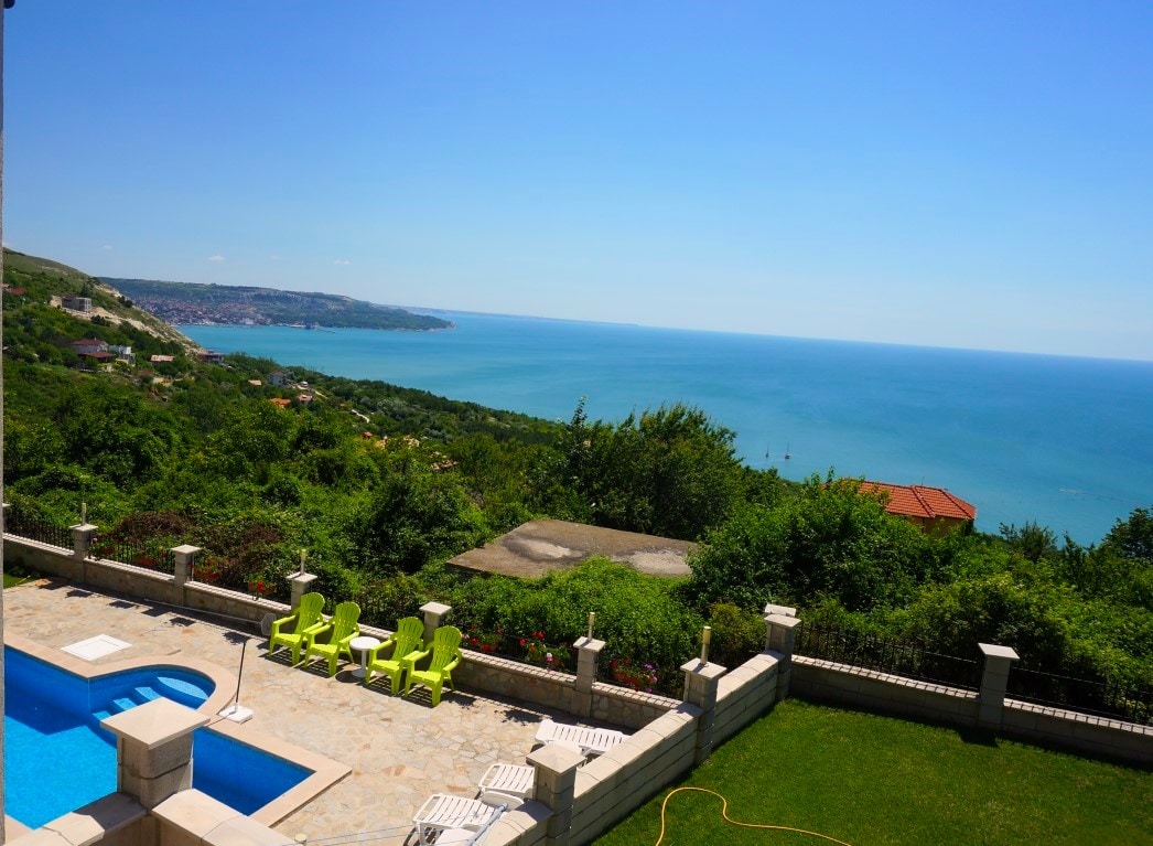 A scenic view showcases a tranquil swimming pool surrounded by a well-maintained garden. Bright green lounge chairs are placed nearby, providing a space to relax. The expansive vista includes the shimmering sea and distant coastal landscape, under a clear blue sky.
