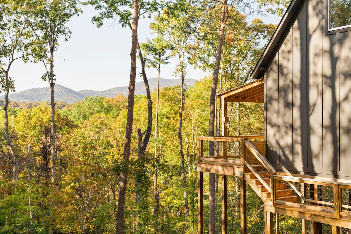 A corner of the cabin is showcased, highlighting its modern architecture amidst a vibrant landscape of autumn foliage. Towering trees surround the cabin, leading to distant mountains that provide a serene backdrop, creating a harmonious connection between the cabin and nature.