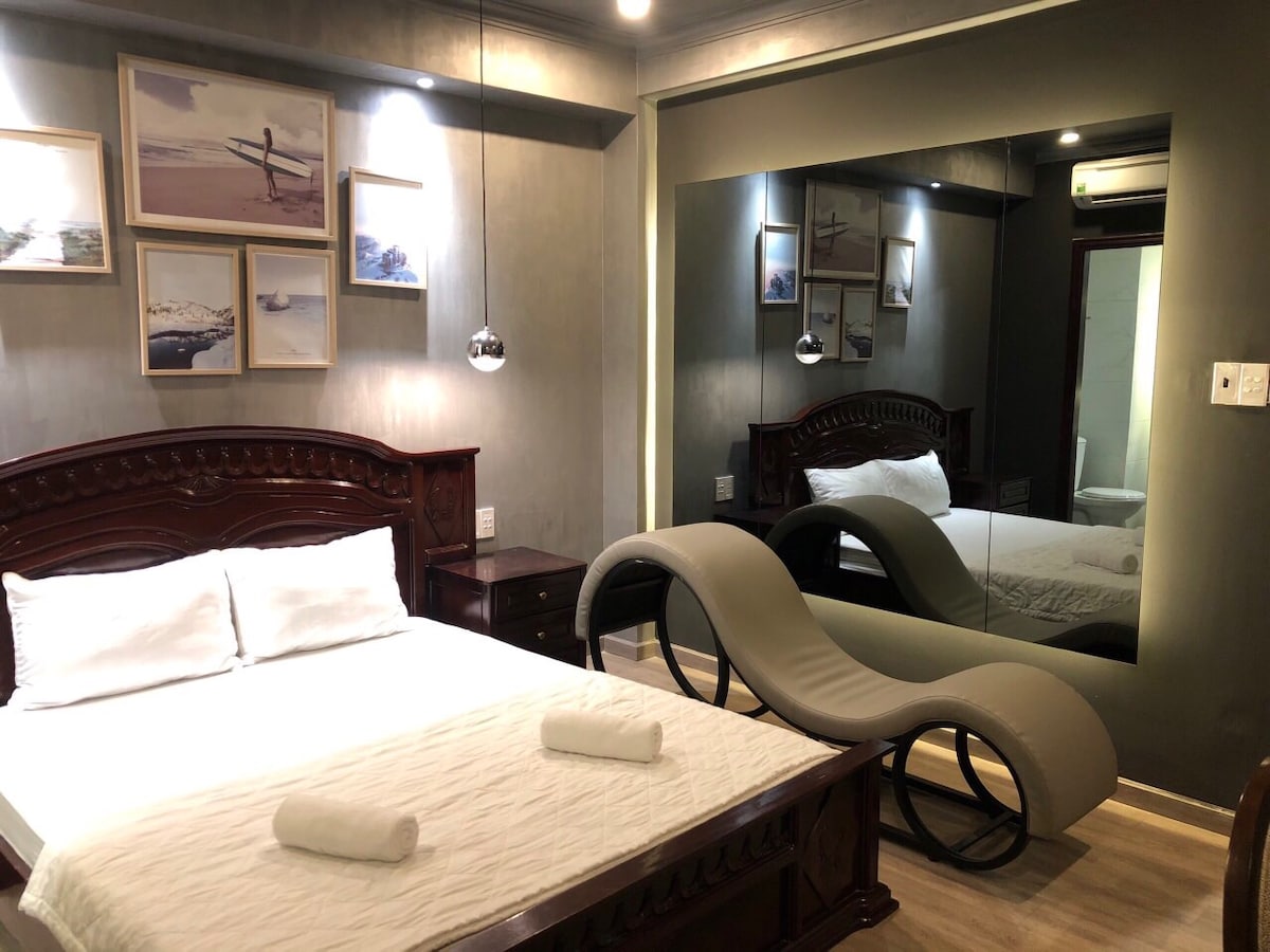 A spacious bedroom features a wooden bed with fresh linens and a pair of neatly rolled towels. A unique, curved lounge chair is positioned beside the bed. The room is complemented by a large mirror, reflecting the decor and providing additional depth.