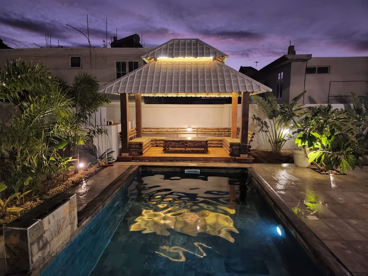 An outdoor pool area features a tranquil swimming pool reflecting the evening sky. A wooden gazebo is positioned nearby, furnished with a seating area made of bamboo. Lush greenery frames the space, while soft lighting illuminates the scene, creating a relaxing atmosphere.