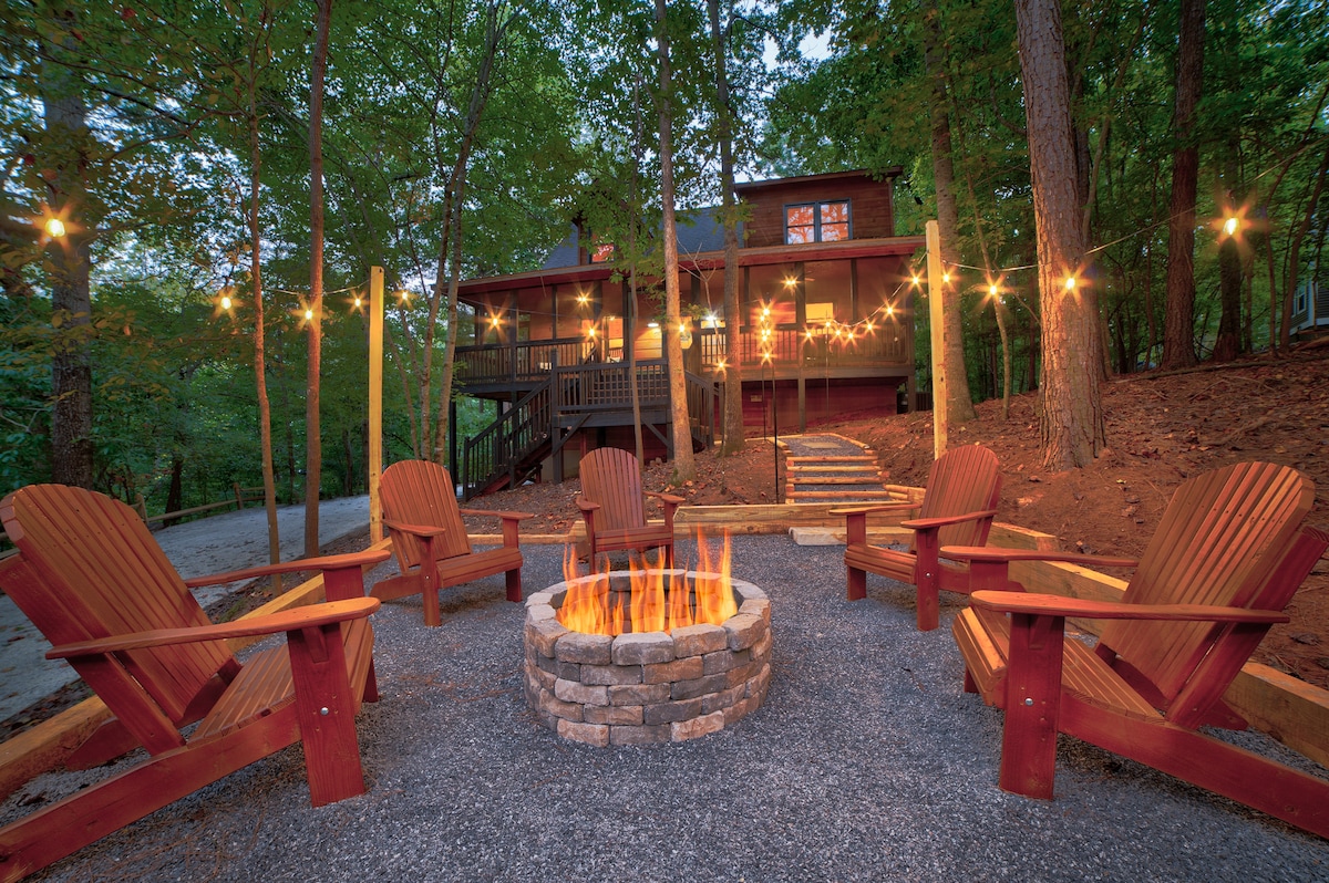 A fire pit surrounded by a stone seating wall is situated in a wooded area, with several wooden Adirondack chairs positioned around it. Soft, warm lights are strung above, creating a cozy atmosphere as the cabin stands in the background, nestled among trees.