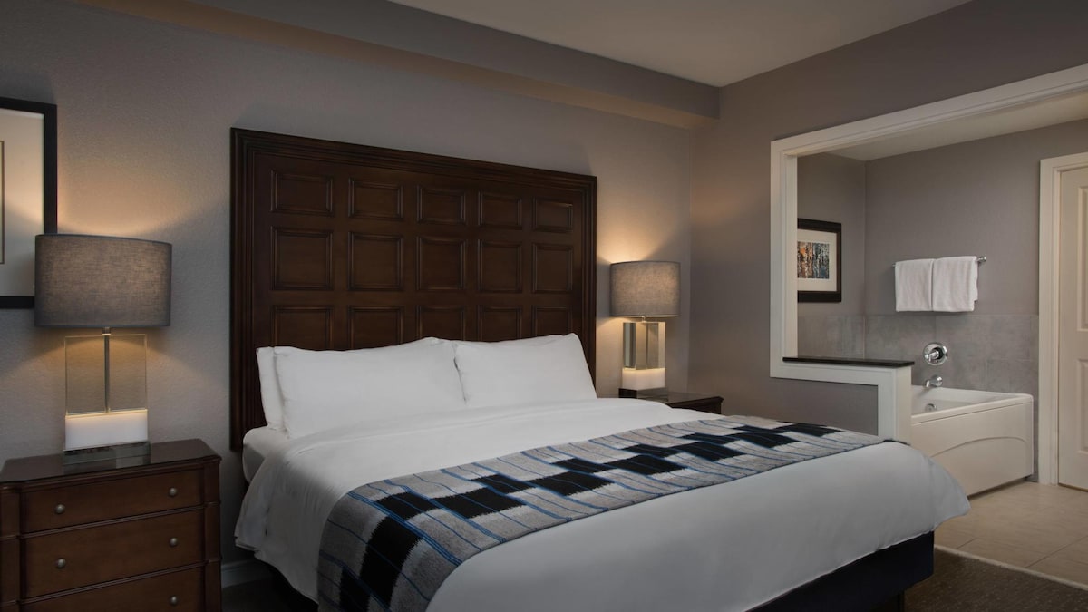 A serene bedroom features a spacious bed with crisp white linens and a decorative blue and black throw. Nightstands with lamps provide soft lighting. The bathroom area is visible in the background, complete with a bathtub and a towel rack.