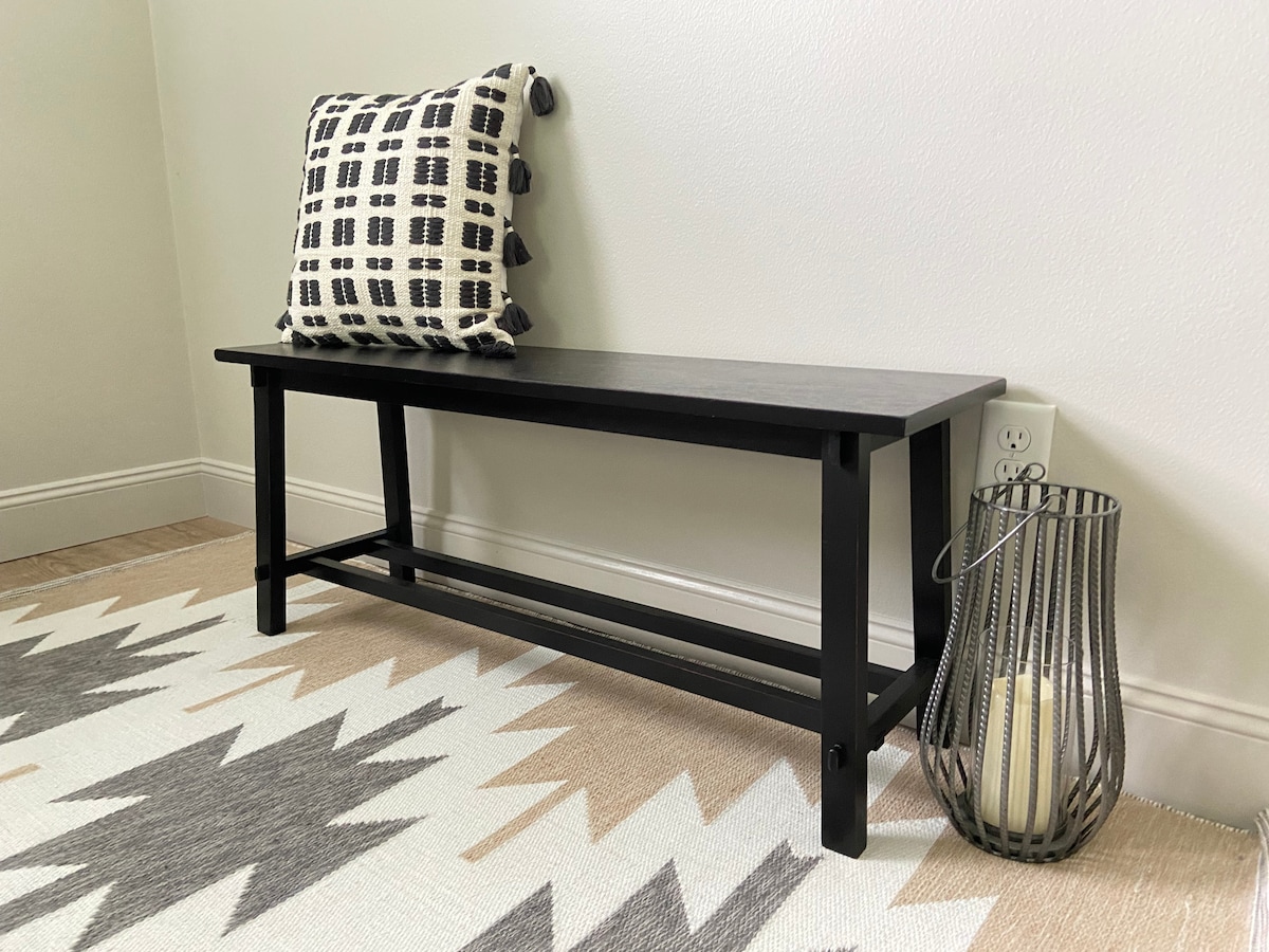 A simple black bench is positioned against a neutral wall, topped with a patterned cushion. A decorative lantern, containing a candle, is placed on the floor beside the bench. A geometric area rug with a mixture of light and dark tones lays beneath.