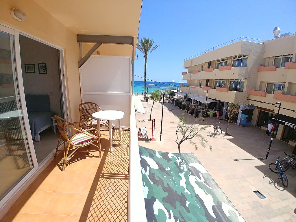 An outdoor terrace features a small round table and two woven chairs, offering a view of a pedestrian square and the beach in the background. Lush palm trees and low-rise buildings are visible, with the sea reflecting a clear blue sky.