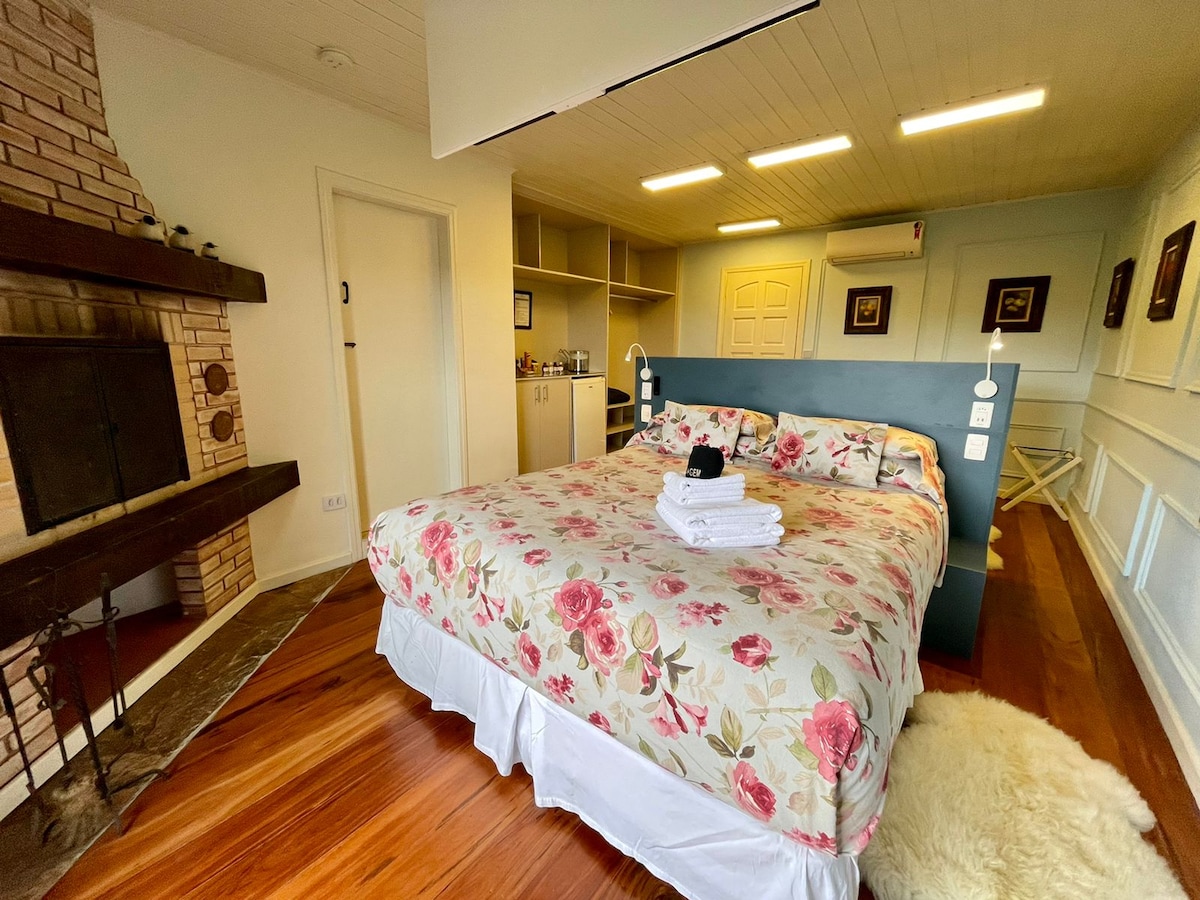 A spacious area features a queen-size bed adorned with floral bedding. A cozy fireplace is situated against the brick wall, while shelves and a small kitchenette are visible in the background. Soft lighting enhances the welcoming environment.