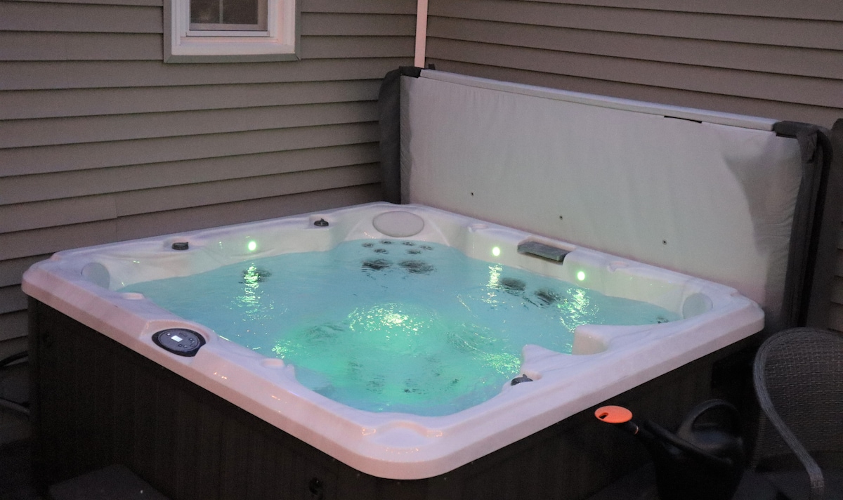 A soothing hot tub is displayed in a private outdoor space, featuring a smooth, white surface and illuminated by soft green lights. The tub is partially covered with a lid, while a chair is positioned nearby.