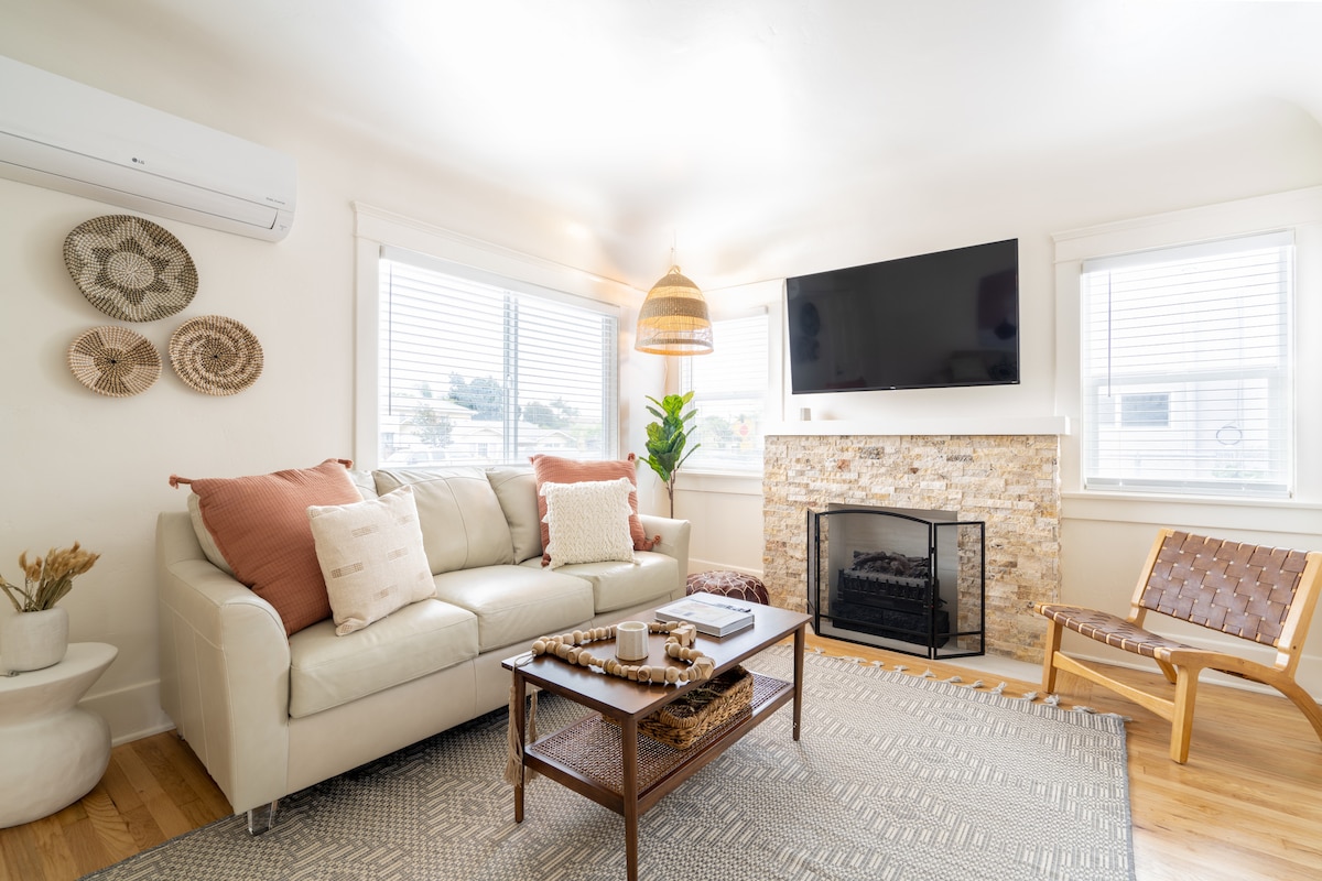 The bright living area features a comfortable light-colored sofa adorned with decorative pillows. A stylish coffee table sits in front, surrounded by a woven area rug. Natural light floods the space through multiple windows, with a cozy fireplace and modern wall-mounted TV adding warmth and entertainment.