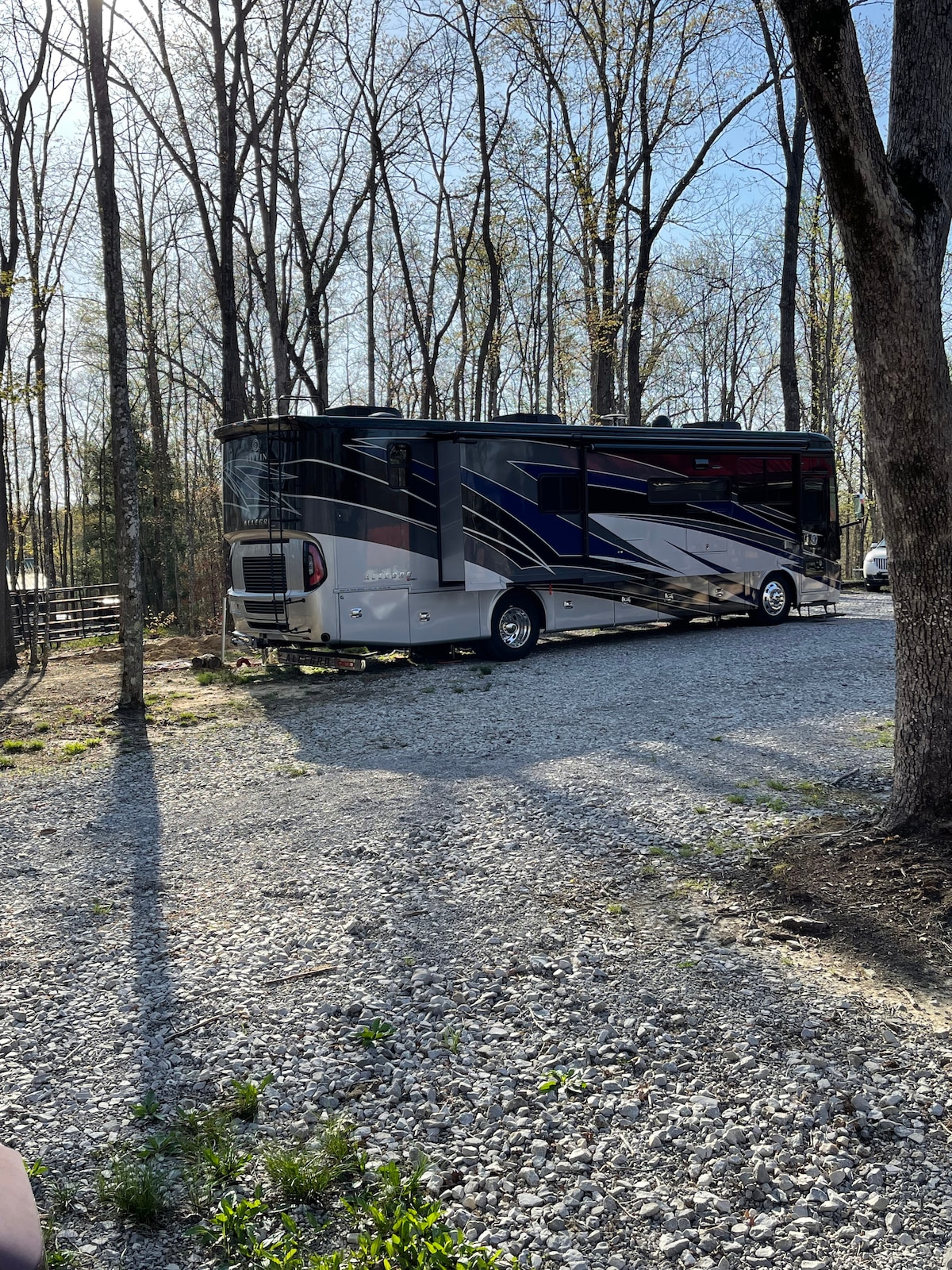 An RV is parked on a gravel surface surrounded by trees, showcasing a well-maintained exterior design. Sunlight filters through the branches, creating dappled shadows on the ground. The spacious area allows for easy maneuverability and access to the campsite.