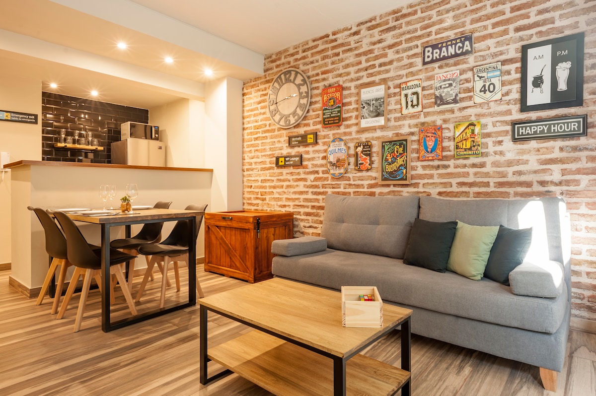 The living area features an industrial design with exposed brick walls, a comfortable sofa, and a wooden coffee table. A dining space is visible, with a set of four black chairs and a wooden table. Decorative wall art adds character to the room.