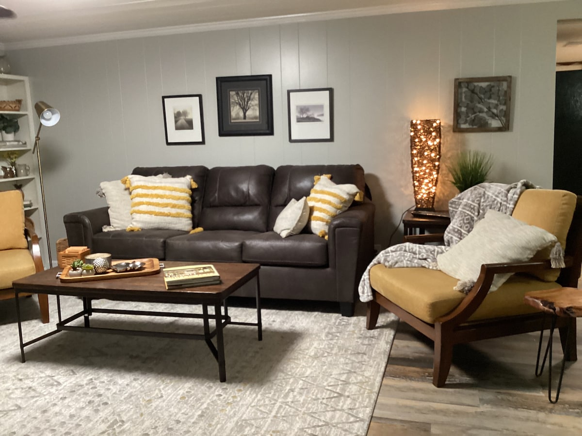 A warm living area is presented with a dark brown sofa featuring decorative pillows. Two armchairs provide additional seating, complemented by a wooden coffee table displaying tasteful decor. Soft lighting from a floor lamp and a textured rug add to the comfort of the space.
