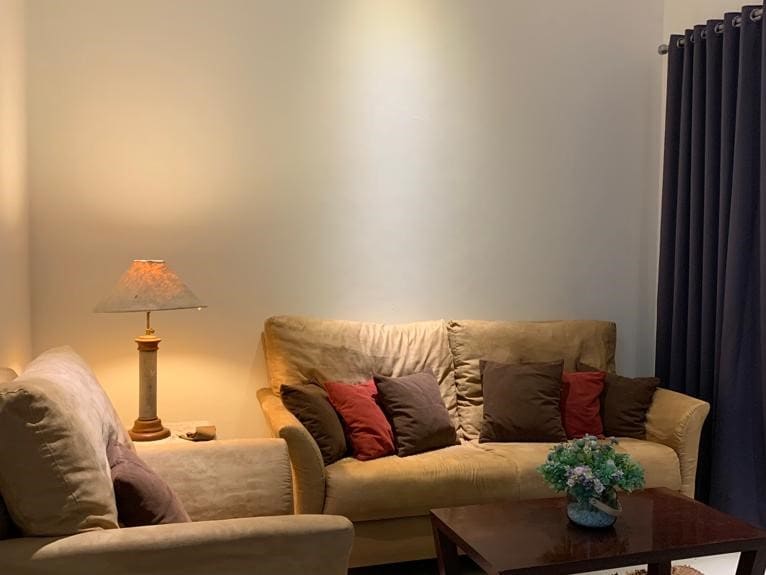 A cozy living area features a light brown sofa adorned with dark and light-colored cushions. A small wooden table sits in front, accompanied by a lamp that softly illuminates the space. Dark curtains frame a window, contributing to a comfortable atmosphere.