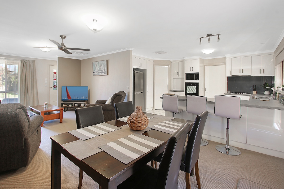 An open plan living, dining, and kitchen area is featured. A dining table with striped placemats sits at the center, surrounded by four upholstered chairs. Modern kitchen appliances are visible, along with a comfortable seating arrangement. Natural light filters through windows, enhancing the spacious feel.