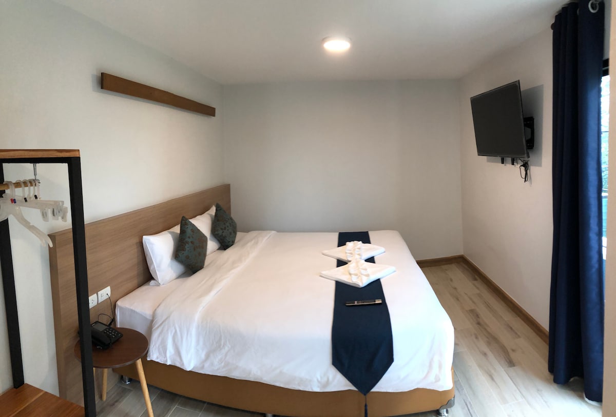A comfortably furnished room features a spacious bed with crisp white linens, complemented by patterned pillows. A flat-screen TV is mounted on the wall, and a seating area with a table can be noticed. Natural light enters through a window, dressed in dark curtains.