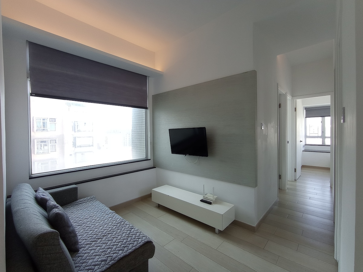 A light-filled living area features a modern gray sofa positioned beside a large window. A flat-screen television is mounted on a pale accent wall, complementing the neutral color scheme. Natural light is filtered through a roll-up shade, creating a serene atmosphere.