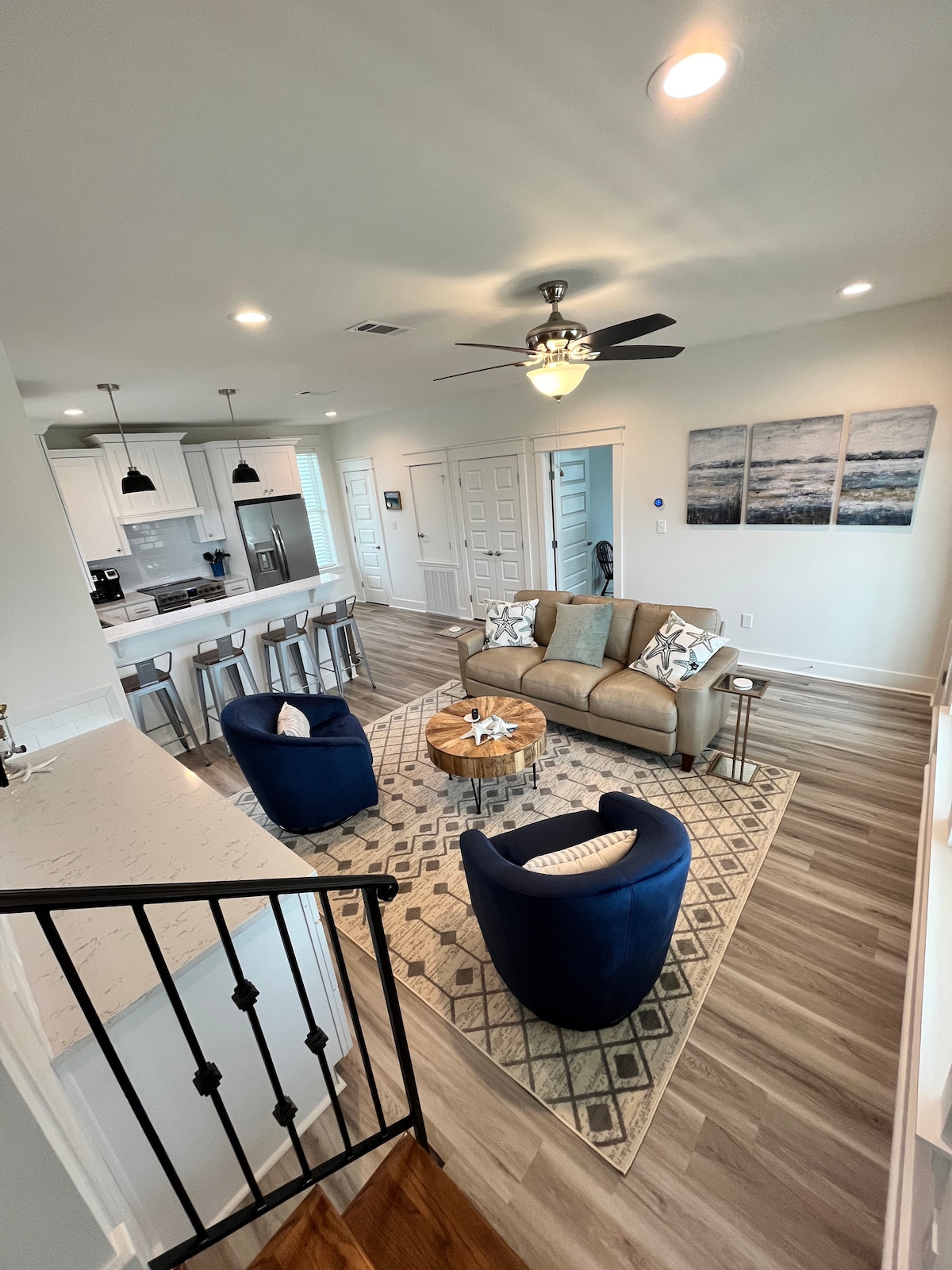 An open-concept living area features a spacious seating arrangement with a beige sofa and two blue chairs surrounding a textured area rug. A round coffee table sits at the center, and light fills the space from overhead fixtures and large windows, creating a welcoming atmosphere.