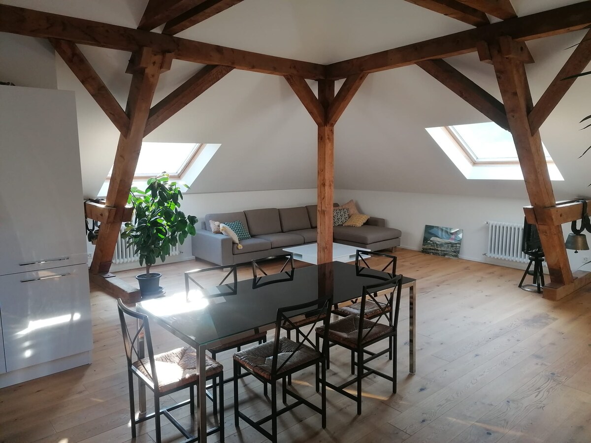 An airy loft space features exposed wooden beams and natural light streaming through skylights. A dining table with chairs occupies the center, while a comfortable sofa is positioned against one wall. The warm wood floor enhances the inviting atmosphere of the room.