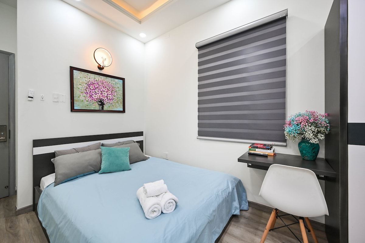 A cozy room features a spacious bed dressed in a light blue blanket, accompanied by pillows and rolled towels. A small desk with a chair is positioned against the wall, alongside a framed artwork of a flowering tree. A window covered by striped shades allows in soft light.