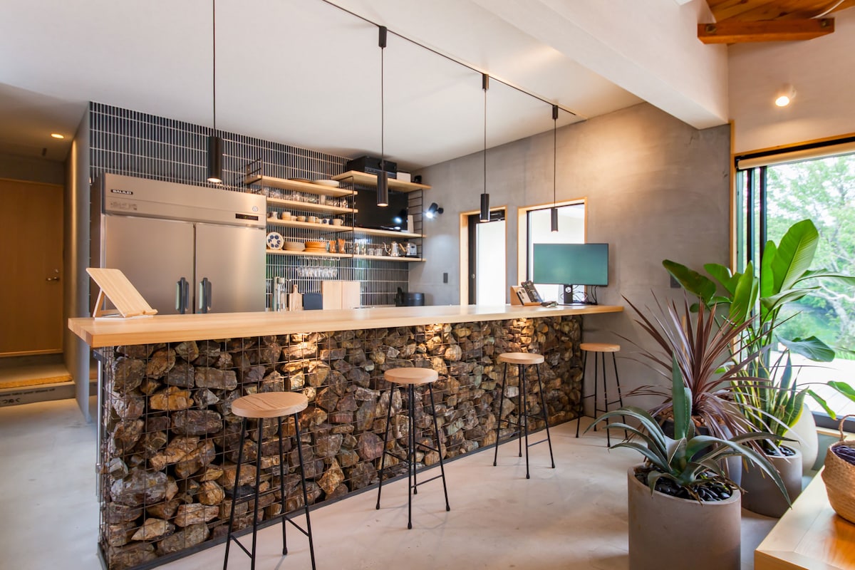 The kitchen area features a modern design with a wooden countertop supported by a stone base. Bar stools are arranged along the counter. Shelving holds kitchen items, while large windows provide natural light and view of greenery.
