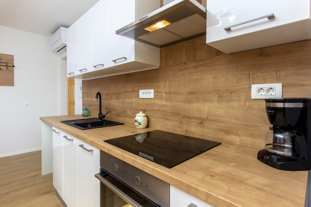 A modern kitchen features sleek white cabinetry paired with a warm wooden backsplash. A built-in stovetop, sink, and coffee maker are visible, reflecting a functional design. Natural light brightens the space, enhancing the inviting atmosphere for meal preparation.