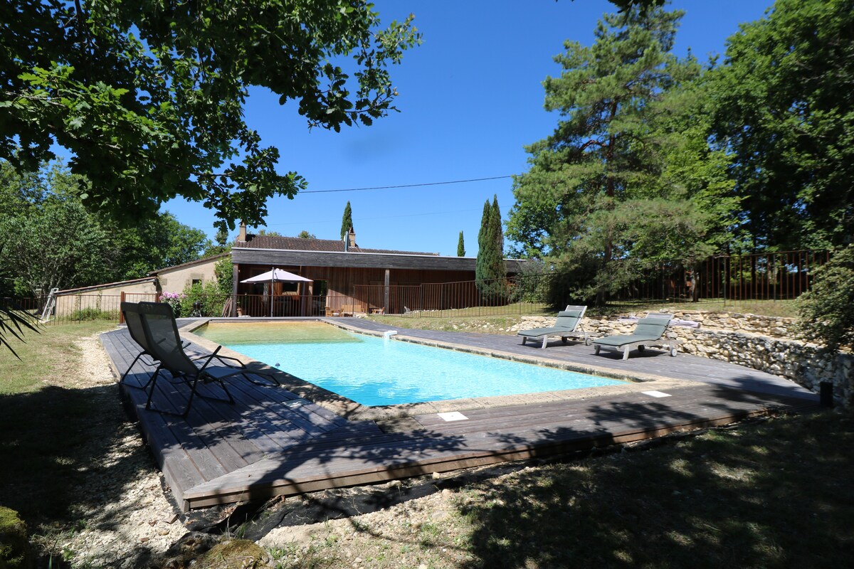A private swimming pool is set within a wooden deck area, surrounded by lush greenery. Sun loungers are positioned along the deck, offering a space for relaxation. The pool is enclosed by a safety fence, ensuring a secure environment for children.