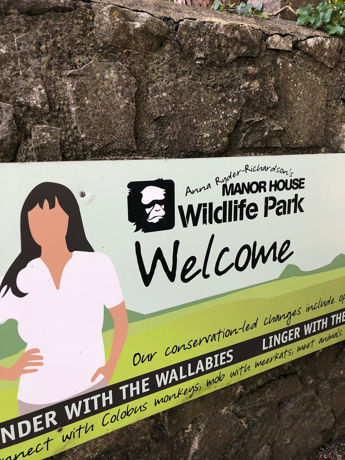 A welcoming sign for Manor House Wildlife Park is displayed, featuring an illustration of a woman in a white shirt. The sign lists various attractions, including interactions with wallabies and colobus monkeys, emphasizing the park's conservation efforts.