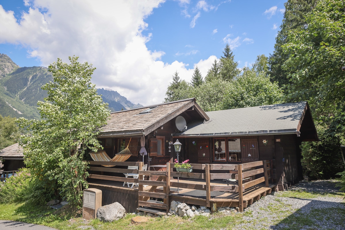 The chalet features a traditional rustic design, surrounded by lush greenery and trees. A wooden deck with railings extends in front, and a covered area is visible. The mountains in the background provide a natural backdrop, creating a serene environment.