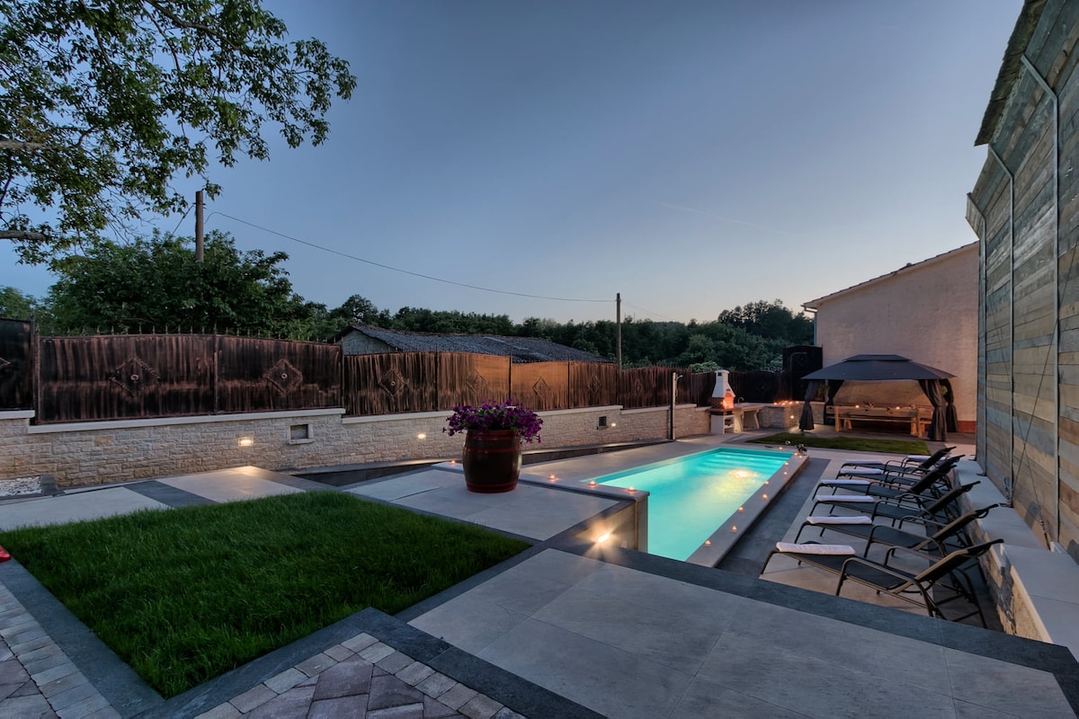 An outdoor space features a rectangular swimming pool illuminated at dusk, surrounded by lounge chairs. A grassy area adds a touch of nature, while potted flowers provide subtle color. A covered seating area can be seen, creating a relaxed atmosphere for outdoor enjoyment.