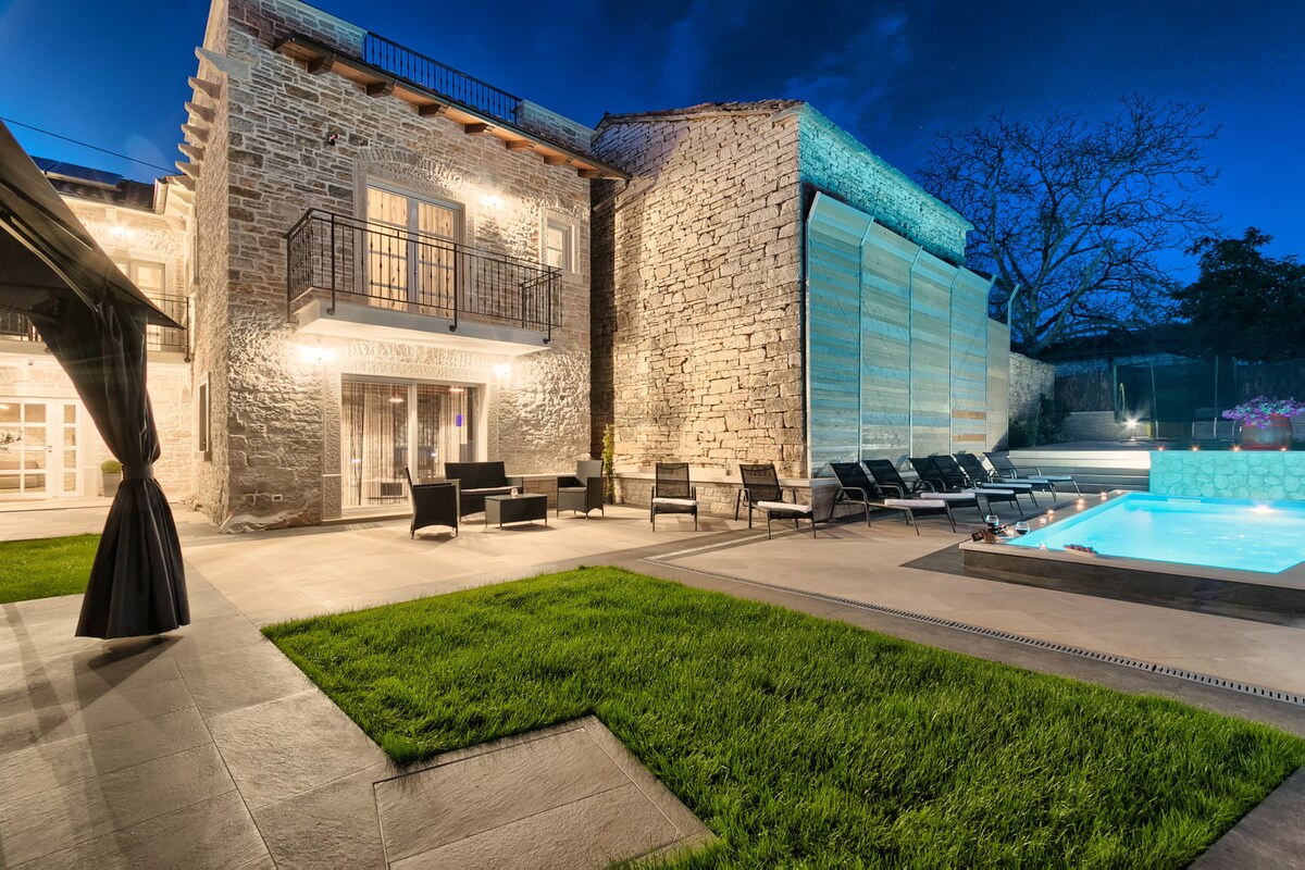 The exterior of Villa Patrick is showcased with the stone facade illuminated at night. A serene pool is visible, surrounded by lounge chairs. The yard features well-maintained grass, and the lighting emphasizes the charm of the villa and the inviting outdoor space.