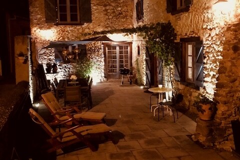 An outdoor patio is illuminated warmly, featuring stone walls and rustic charm. Comfortable seating options are arranged, including wooden lounge chairs and a dining area. Potted plants and a grill are also present, creating a welcoming space for relaxation or gatherings.