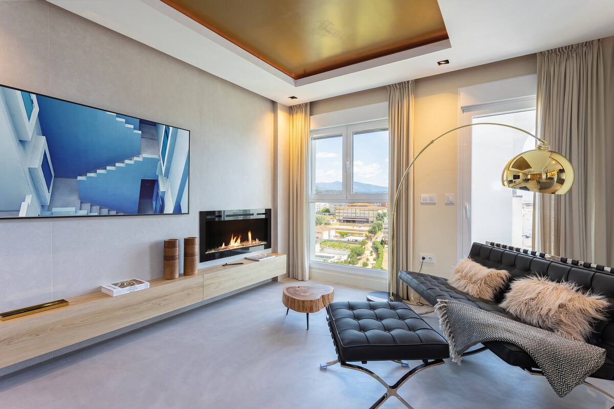 A modern living area features a sleek black lounge chair and an elegant curved floor lamp. A flat-screen TV is mounted on a wooden media unit, while a fireplace adds a cozy touch. Large windows offer views of the park and nearby surroundings.