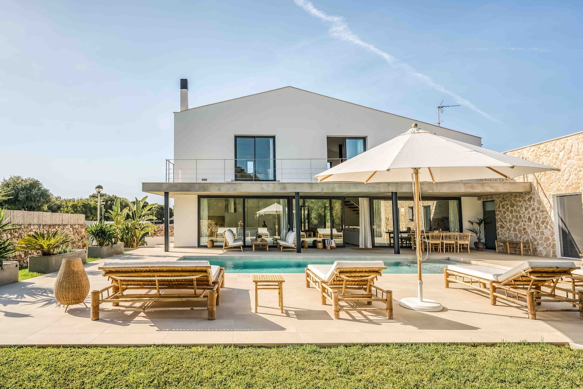 A modern villa features a spacious outdoor area with comfortable wooden lounge chairs surrounding a turquoise pool. An umbrella provides shade over a seating area. Large glass doors offer a view of the interior, blending outdoor and indoor living seamlessly.